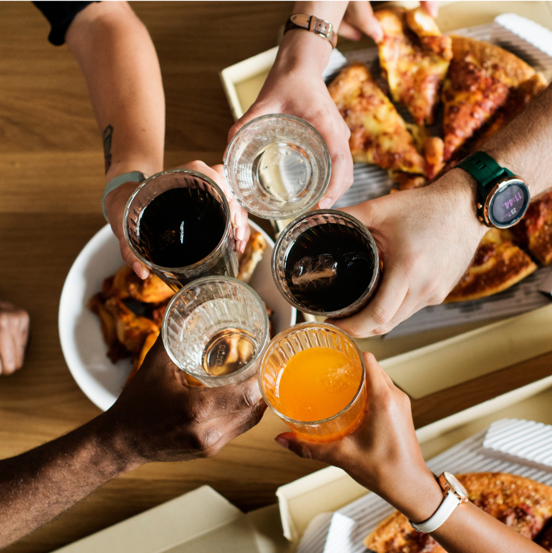 Five hands holding glasses full of liquid and touching them together over a table full of food